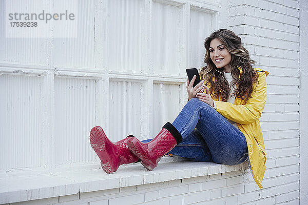 Smiling woman sitting white wall using mobile phone