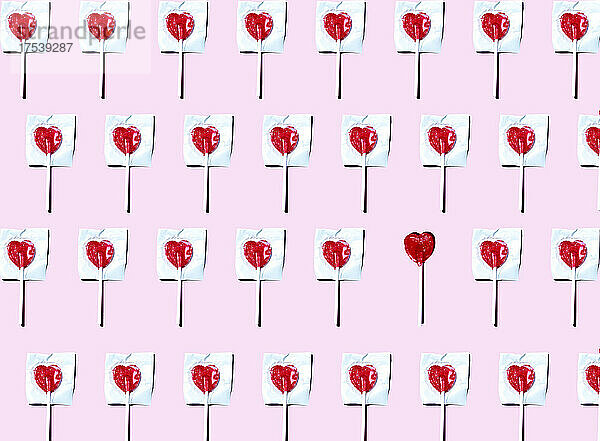Pattern of heart shaped lollipops flat laid against pink background with single one unwrapped