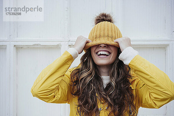 Playful woman covering face with knit hat in front of wall