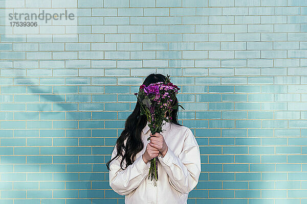 Young woman hiding face behind bouquet in front of brick wall