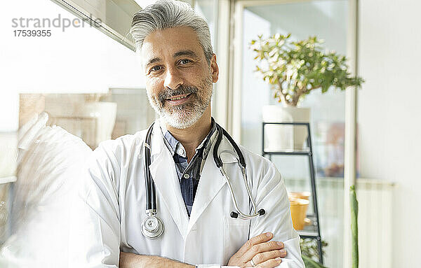 Smiling doctor with arms crossed leaning on window at home