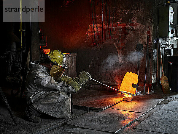 Handwerker trägt Schutzkleidung und gießt Gussmetall in ein Stahlwerk
