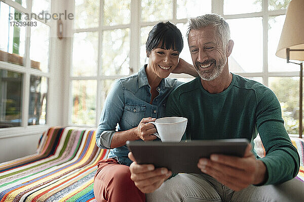 Smiling man sharing tablet PC with woman holding coffee cup at home