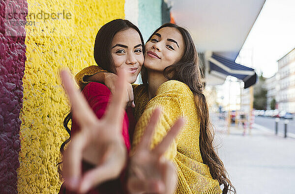 Young sisters embracing and gesturing with peace sign by colorful wall