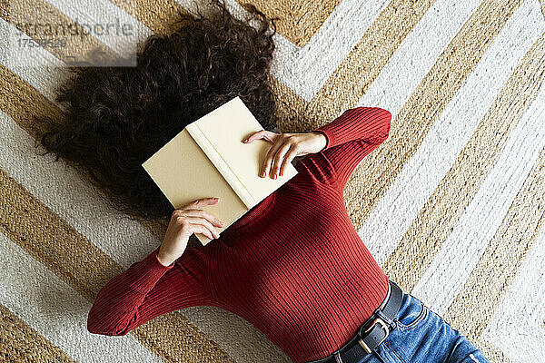 Woman covering face with book lying on carpet at home