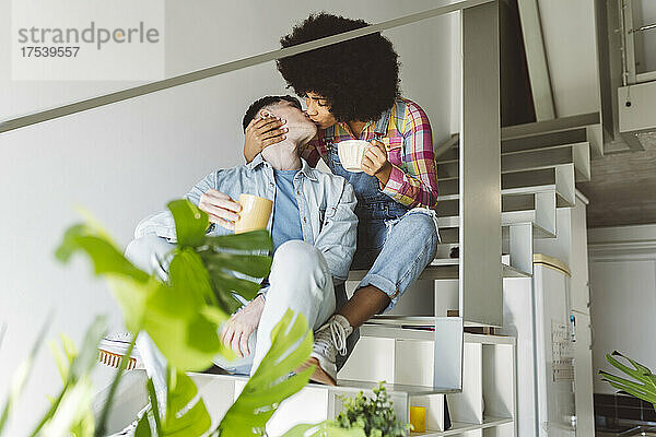 Young couple holding coffee mugs kissing on staircase at home