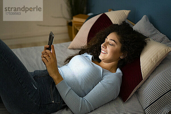 Smiling woman using smart phone lying on bed at home