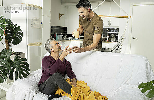 Lächelnder schwuler Mann gibt seinem Mann  der zu Hause auf dem Sofa im Wohnzimmer sitzt  eine Kaffeetasse