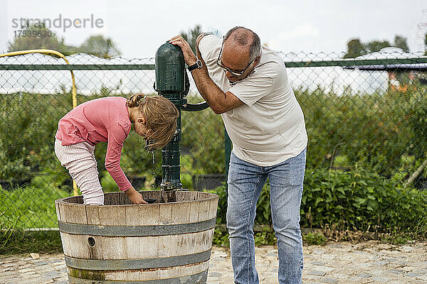 Grandfather pumping water in barrel with granddaughter inside