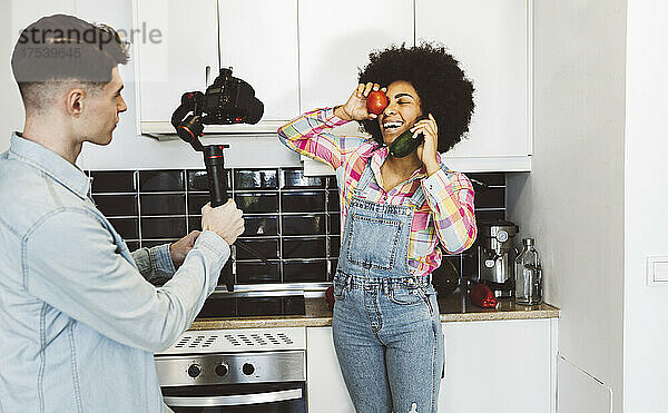Young man filming playful woman holding vegetables in kitchen