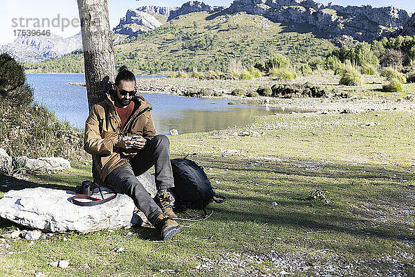 Wanderer sitzt mit Mobiltelefon auf einem Felsen am Cuber-Staudamm  Mallorca  Balearen  Spanien