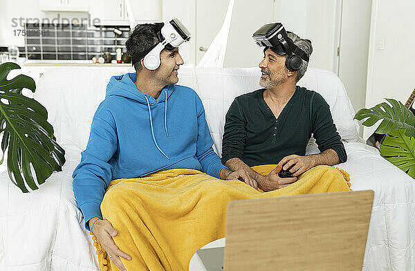 Smiling gay couple wearing VR glasses looking at each other sitting on sofa