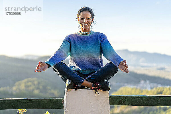 Smiling woman sitting in lotus position on railing at park