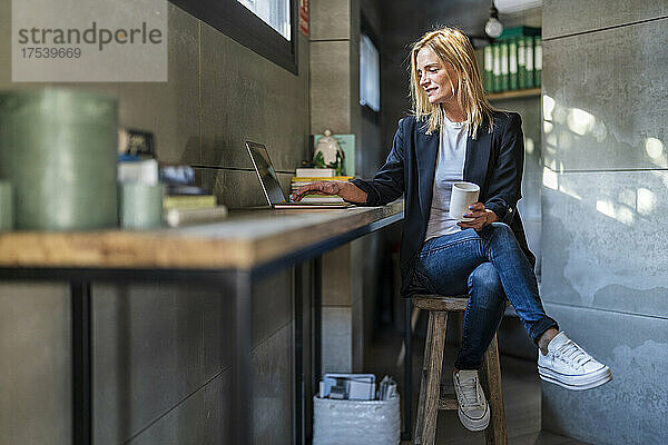 Geschäftsfrau mit Kaffeetasse und Laptop sitzt auf einem Hocker im Büro