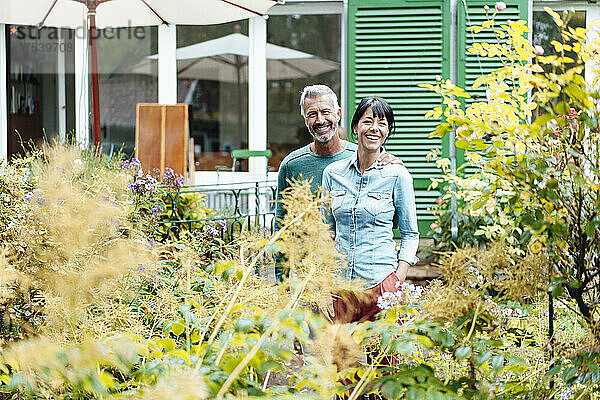 Happy man arm around woman amidst plants in backyard