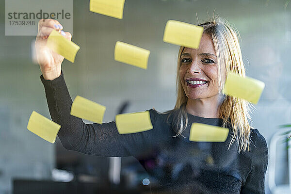 Smiling businesswoman writing on adhesive notes seen through glass wall at office