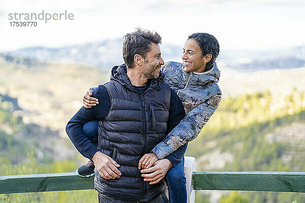 Smiling woman sitting on railing hugging man from behind at park