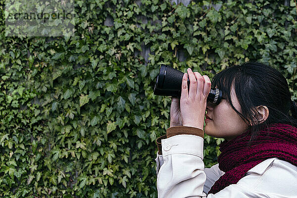 Woman using binoculars near green ivy wall