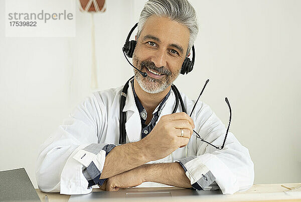 Smiling doctor with headphones holding eyeglasses at home office