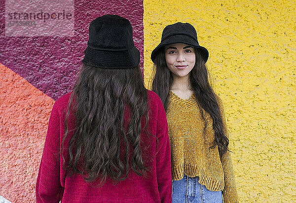 Smiling young woman standing by sister facing colorful wall