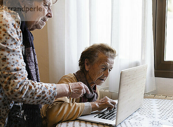 Senior woman assisting elderly sister using laptop at home