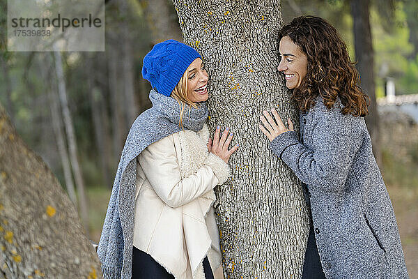 Cheerful friends looking at each other leaning on tree trunk