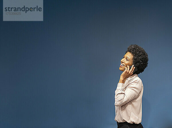 Happy businesswoman talking on smart phone by blue wall in office