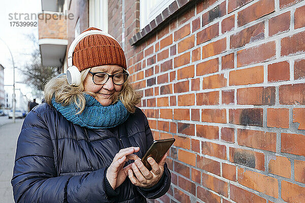 Senior woman with headphones text messaging on mobile phone by brick wall