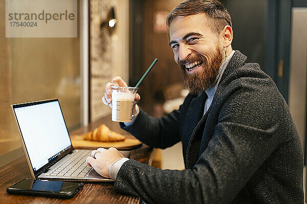 Fröhlicher Geschäftsmann mit Kaffeetasse sitzt mit Laptop und Smartphone im Restaurant