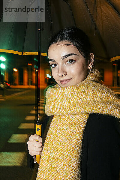 Smiling teenage girl with scarf and umbrella on street at night