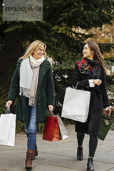 Lächelnde Frauen mit Einkaufstüten schauen sich beim Gehen auf dem Fußweg an