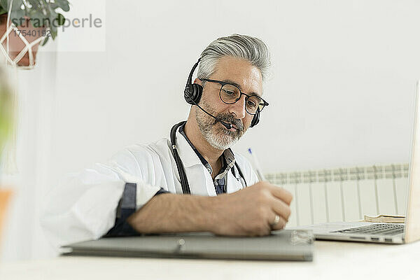 Arzt mit Kopfhörern schreibt im Heimbüro auf die Zwischenablage