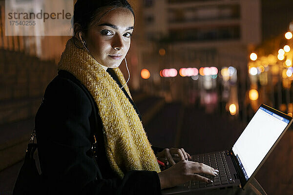 Teenage girl with laptop listening music on in-ear headphones at night
