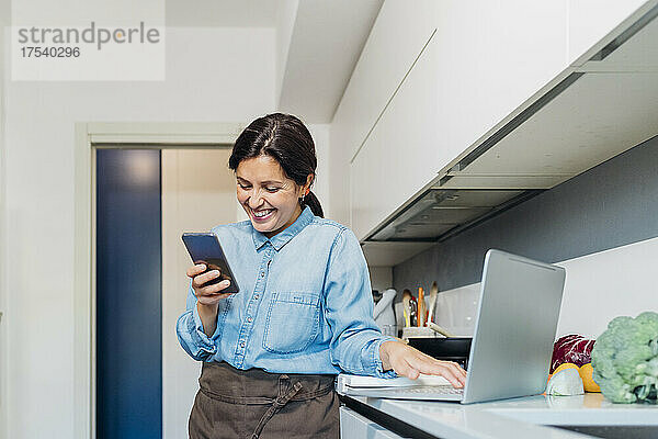 Smiling woman using mobile phone and laptop standing by kitchen counter at home