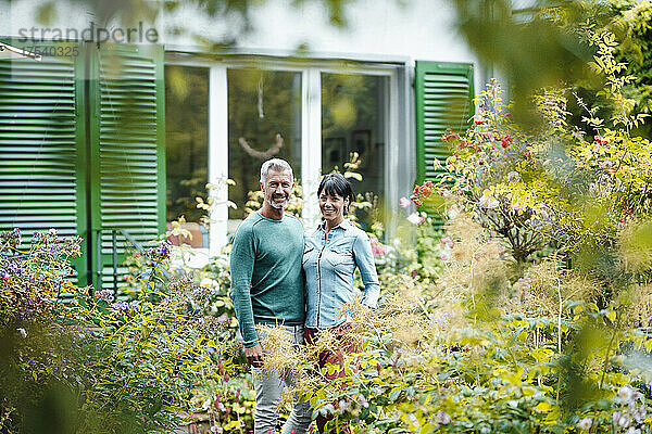 Smiling mature couple standing in backyard seen through plants