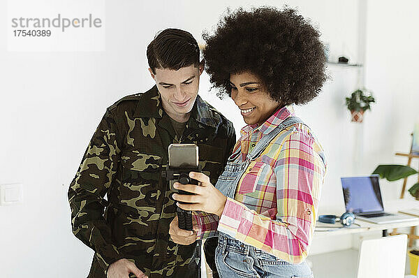 Smiling woman sharing smart phone with young soldier man at home