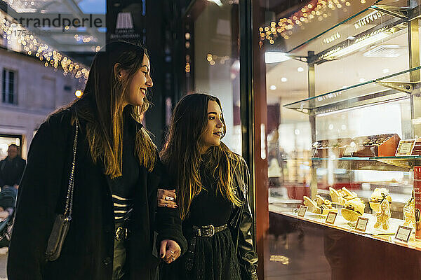 Smiling friends looking at food on display through illuminated store window