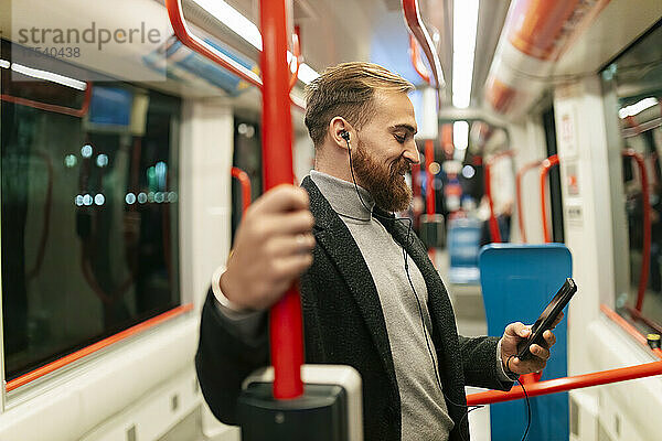 Man standing in tram using smart phone and listening music through in-ear headphones