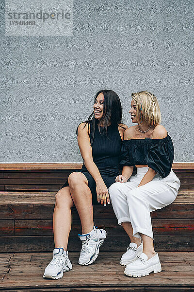 Smiling women sitting together on bench in front of wall
