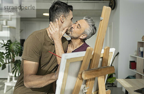 Affectionate gay couple kissing by canvas at home