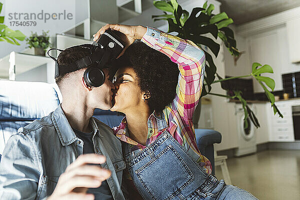 Romantic man kissing young woman holding VR headset in living room