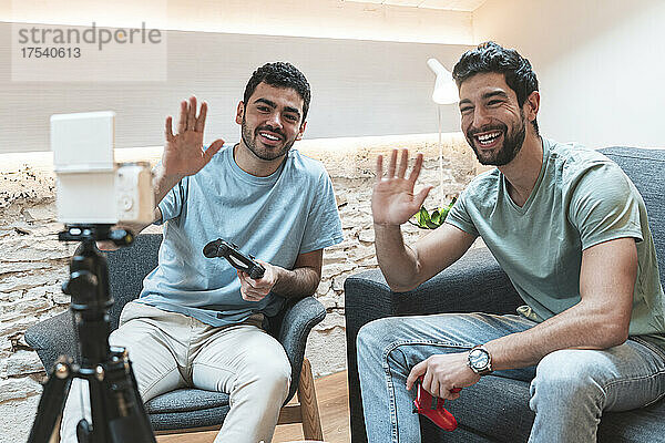 Lächelnde Freunde mit Videospiel-Controllern winken auf einem Stativ in die Kamera