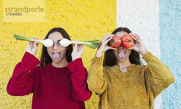Playful sisters covering eyes with vegetables in front of multi colored wall
