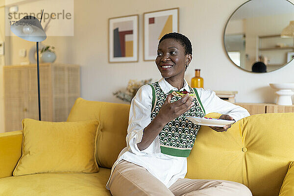 Smiling woman having breakfast on yellow sofa at home