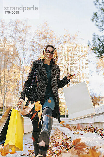 Woman kicking autumn leaves walking on footpath holding shopping bags