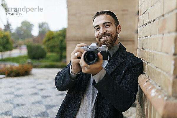 Happy young bearded man with camera leaning on wall