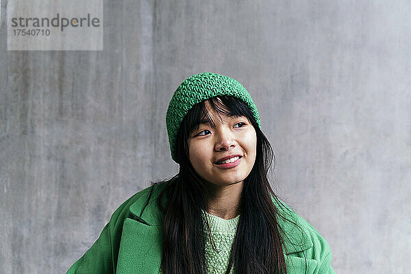 Smiling woman wearing green knit hat day dreaming in front of wall