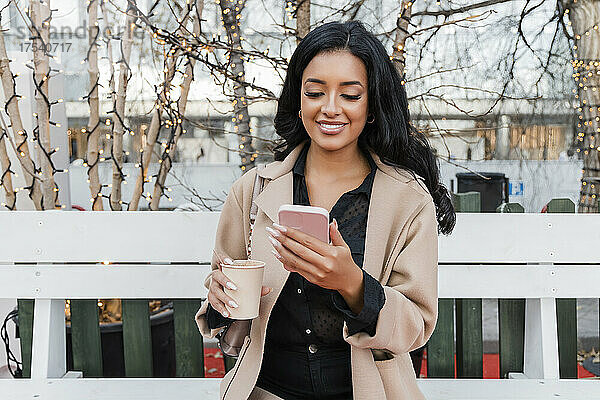 Lächelnde Frau mit Kaffeetasse und Smartphone auf der Bank