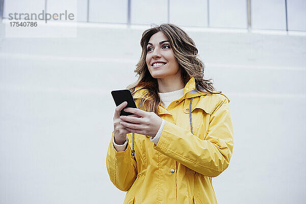 Smiling young woman holding mobile phone in front of wall