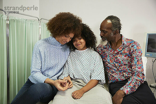 Frau umarmt Tochter  die neben Vater im Krankenhausbett sitzt